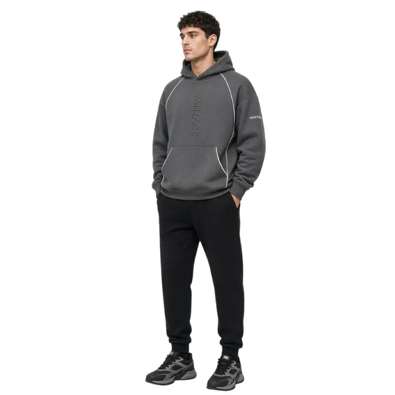 A man wearing a dark gray pullover hoodie with white piping details and black sweatpants, standing against a plain white background.
